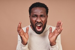 Angry young African man in casual clothing screaming and looking at camera while standing against brown background
