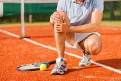 Sports injury. Close-up of tennis player touching his knee while sitting on the tennis court