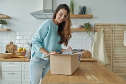 Beautiful young woman unpacking box while standing at the domestic kitchen
