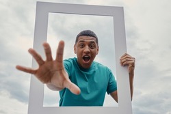 Playful mixed race man looking through a picture frame while standing outdoors