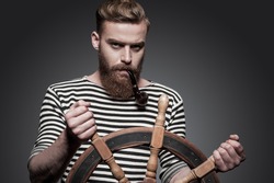 Finding the right way. Confident young bearded sailor steering a wheel while standing against grey background 