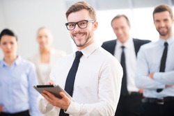 Confident business expert. Confident young businessman holding digital tablet and smiling while his colleagues standing in the background 