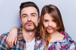 Funny moustache. Beautiful young loving couple making fake moustache from hair while standing against grey background