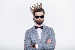 King of style. Sneering young handsome man wearing suit and crown keeping arms crossed and looking at camera while standing against white background