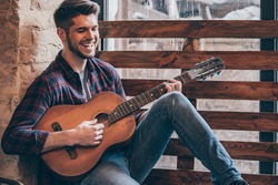 Cheerful guitarist. Cheerful handsome young man playing guitar and smiling while sitting at windowsill 