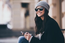 What a beautiful day! Side view of beautiful young woman in sunglasses using her smartphone and looking away with smile while sitting outdoors