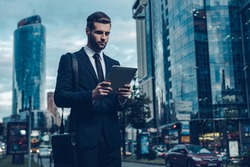 Modern businessman. Night time image of confident young man in full suit holding digital tablet and looking at it while standing outdoors with cityscape in the background