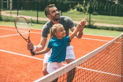 Tennis is fun when father is near. Cheerful father in sports clothing teaching his daughter to play tennis while both standing on tennis court 