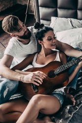 Beautiful couple playing guitar. Handsome young bearded man teaching his girlfriend to play guitar while both sitting in bed at home together