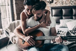 Playing by heart. Handsome young bearded man sitting in bed and playing guitar while attractive woman embracing him 