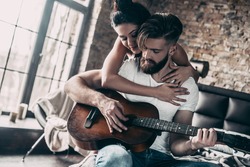 Guitar fun. Handsome young bearded man sitting in bed at home and playing guitar while attractive woman embracing him