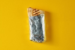 Large shawarma with meat and vegetables, wrapped in foil on a yellow background. Close-up, selective focus
