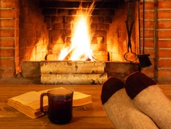 Cup of tea, book, women's feet in warm socks on a wooden table opposite a burning fireplace