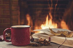 Red mug with hot tea and an open book in front of a burning fireplace, comfort, relaxation and warmth of the hearth concept.