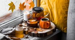Autumn still life with beautiful bokeh. A Cup of hot tea, a teapot, honey, candles and a knitted blanket on the windowsill. Outside the window, autumn leaves and raindrops.