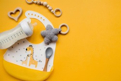 Serving baby food. Colorful silicone bib with giraffes, spoon and milk bottle on a trendy yellow background, flat lay. 
