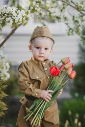 9th May. A little boy 2 years old in military uniform with a bouquet of red tulips on a background of cherry blossoms.The concept of the Great Victory Day 1941-1945 and Defender of the Fatherland Day.