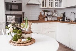 An iron pot with Easter eggs, flowers and rabbits on the table.In the background is a white Scandinavian-style kitchen. The concept of home comfort and decor in the bright holiday of Easter 2022.
