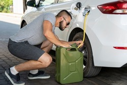 A young man steals gasoline from the gas tank in the white car. A young man pumps gasoline from a gas tank into a canister. Fuel and oil crisis. The concept of gasoline prices and the oil crisis.