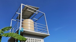 Water tank on the tower with solar panels. Close up water tank with solar panels on top for pumping soil in garden or farm water on blue sky background with copy space. Selective focus