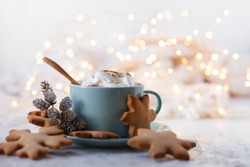 Hot winter drink: chocolate with whipped cream in blue mug. Christmas time. Cozy home atmosphere, white background. Homemade gingerbread cookies, cones and lights as decor. Holiday mood in the air!