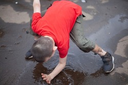 Child falls into puddle. Boy gets his hand dirty in dirty water. Child outside in summer. Schoolboy in red T-shirt. Troubled teenager hits ground.