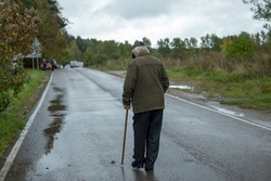 Old man walks down road. Pensioner with stick for support. Man with leg injury. Grandpa in Russia walks through park.