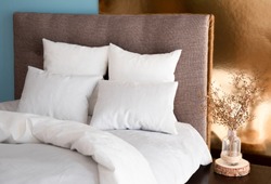White pillows, duvet and duvet case on a bed with brown headboard. White bed linen on the sofa. Bedroom with bed, white bedding, nice posy on the bedside and colored wall. Right side view.