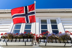 Norwegian national flag flying proudly in front of a traditional white wooden house with a  window box of flowers and blue sky above on a perfect Summer day, Stavanger, Norway
