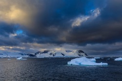 Dramatic sky with low cloud illuminated by storm light over awe inspiring frozen blue Antarctic scenery of icebergs, glaciers and mountains 