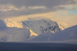 Antarctic sunrise causes snowy spectacular mountains to glow with golden light in an atmospheric scenic view of the Antarctic scenery of the glacial coast of the icy Antarctic Peninsular of Antarctica