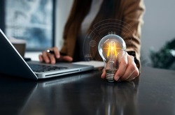 Businesswoman hand holding lightbulb and using laptop computer in office, Idea saving energy and business technology concept.