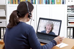 Woman with headset at her desk in front of her laptop having a video chat with her happy grandmother, text space