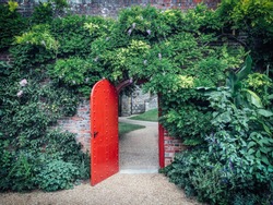 Open, red, half-round door in a brick wall covered with green vines, behind a door a winding gravel path. Lush green foliage cover the brick wall. A mysterious path leads towards the castle walls.