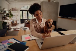 Happy african woman working at home using laptop during quarantine sitting with dog