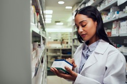 Young female pharmacist reading instructions on medicine box standing in aisle of chemist