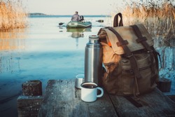 Enameled mug of coffee or tea, backpack of traveller and thermos on wooden pier on tranquil lake. A fisherman on rubber boat in background.