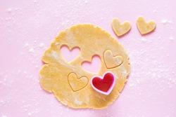 Cutting out heart-shaped cookies from rolled dough with a pink plastic mold. Top view on flour sprinkled gently pink background.