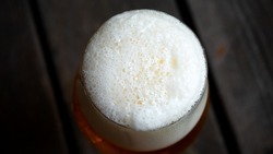 Foamy lager beer, captured from above. The white bubbly foam fills the glass to the lip. Wooden boards fills the background. Beer consumption and brewing concept.