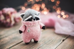 Handmade knitted toy. Amigurumi. Raccoon toy in the gentle pink jumpsuit on the wooden background. Crochet stuffed animals. 