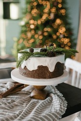 Festive Christmas cake with white icing decorated with spruce branches stands on a set table against a background of lights