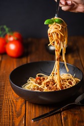 A hand holding a fork with spaghetti with bolognese tomato sauce in black bowl on wooden table