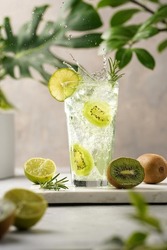 A green refreshing beverage with kiwi, lime and ice cubes in tall long glass with splashes on marble board in green plants setting