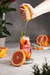 Female hand squeezing juice from orange into a tall glass with cold orange juice with grapefruit, frozen berries and ice cubes, rosemary, grapefruit half on grey background