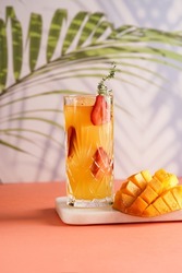 tall cocktail glass filled with yellow juice and fresh strawberries and cut in cubes mango half on tropical palm tree background