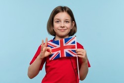 Nice girl 9-11 y.o. wearing basic red t-shirt and blue headphones on light blue background in studio looking at camera. Child holding UK flag. Concept of Studying in United Kingdom of Great Britain.