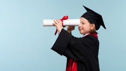 Funny Whizz kid girl wearing graduation cap and ceremony robe on light blue background. Graduate celebrating graduation. Education Concept. Smart child looks to the future through the certificate
