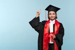 Proud child girl in graduate suit with certificate diploma on light blue background looking at camera, showing promo, pointing finger up, inviting use link. Graduate Celebrating Graduation.