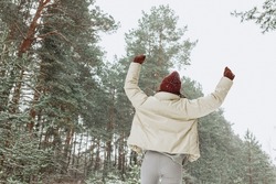 Back view of carefree traveling woman in outerwear enjoying freedom and celebrating success with raised hands in snowy forest in winter 