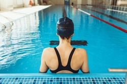 Back view of professional female swimmer in cap and swimsuit standing on lane in pool during training 